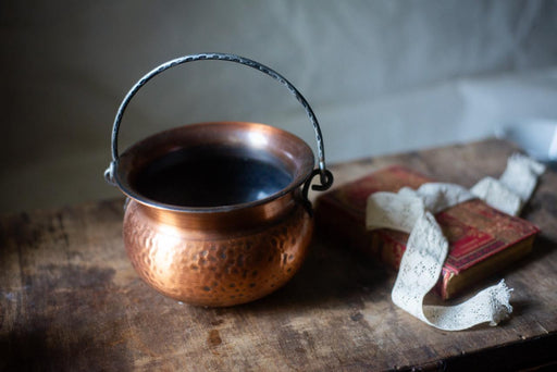 A copper-colored pot with a metal handle sits on a wooden surface, accompanied by a white lace ribbon and a red book.