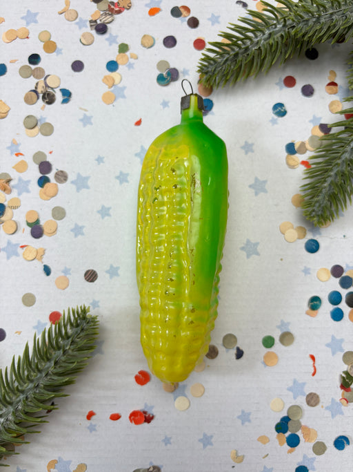 A green and yellow corn cob ornament is placed on a white background with scattered confetti and pine branches.