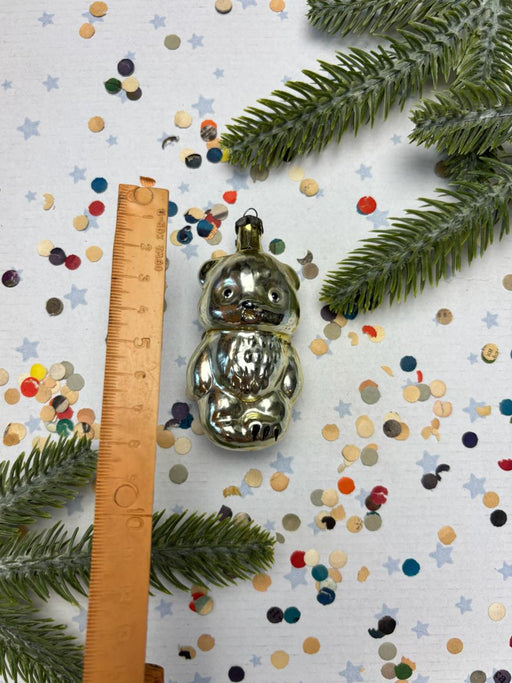 A shiny metal ornament in the shape of a bear is placed next to a ruler on a background of confetti and pine branches.