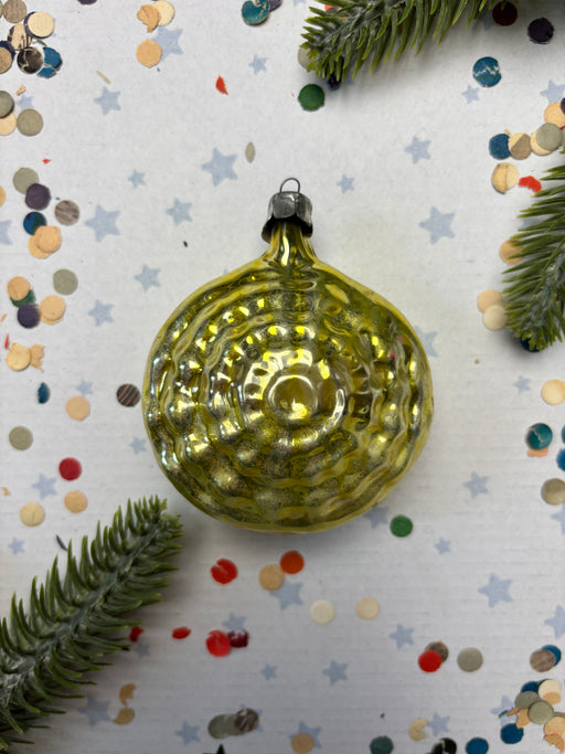 A shiny, golden Christmas ornament is placed on a white background, surrounded by scattered confetti and pine branches.