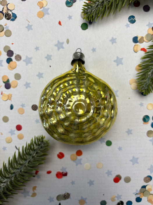 A shiny, golden Christmas ornament is placed on a white background, surrounded by scattered confetti and pine branches.