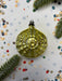 A shiny, golden Christmas ornament is placed on a white background, surrounded by scattered confetti and pine branches.