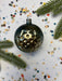 A shiny, metallic ornament with a star-like design sits on a white background, surrounded by scattered confetti and a sprig of greenery.
