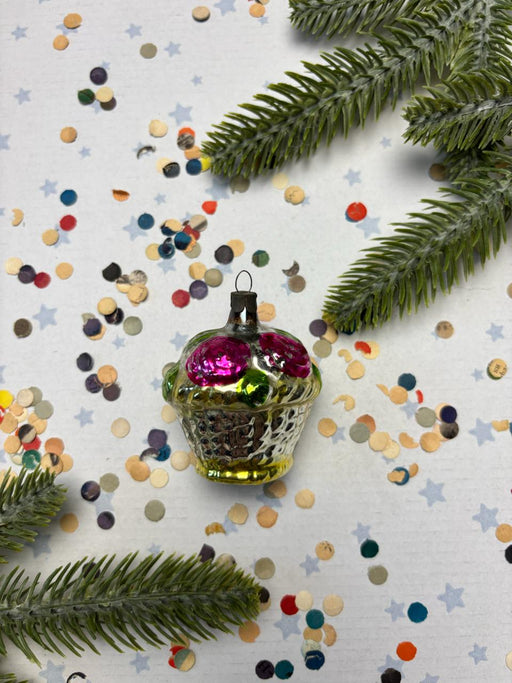 A glass ornament with a pink flower is surrounded by colorful confetti and a pine branch on a white background.