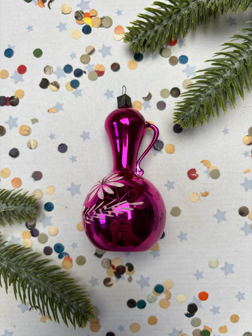 A pink glass ornament with a handle and a flower design is placed on a white background, surrounded by scattered confetti and green pine branches.