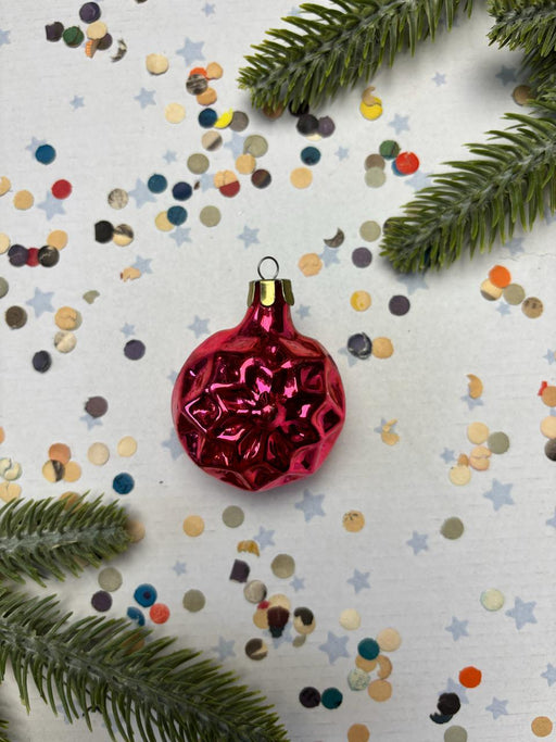 A red Christmas ornament is surrounded by colorful confetti and green pine branches on a white background.