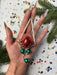 A hand holding a red ornament with green and silver accents, surrounded by Christmas decorations and confetti.