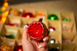A hand holding a red ornament in front of a blurred background filled with various Christmas ornaments.