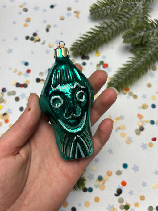 A hand is holding a green ornament with a face design, surrounded by confetti and a pine branch.