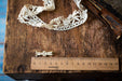 a wooden ruler with a scale ranging from 0 to 15 centimeters, placed on a wooden surface alongside a piece of white lace and a small golden pin.