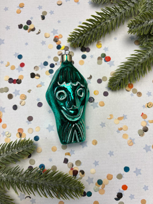 A green ornament with a face design is surrounded by colorful confetti and pine branches on a white background.