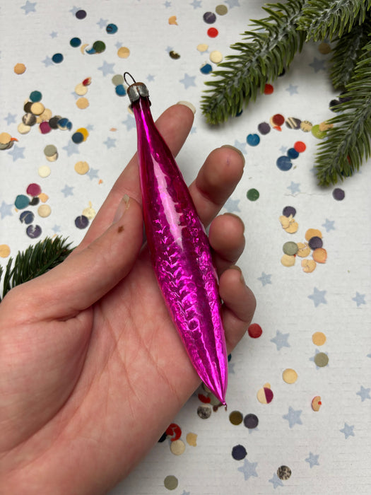 A hand holding a pink ornament against a background of colorful confetti and a pine tree branch.
