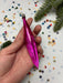 A hand holding a pink ornament against a background of colorful confetti and a pine tree branch.