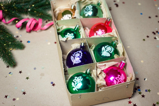 A cardboard box containing a variety of colorful Christmas ornaments, including green, purple, and pink ornaments, along with some confetti on the ground.