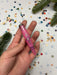 A hand holding a pink crayon with a floral design, surrounded by colorful confetti on a white background.