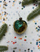 a Christmas ornament with a green leaf on it, surrounded by confetti and pine branches.