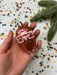 A hand holding a small, orange ornament with a red and white design, surrounded by colorful confetti on a white background.
