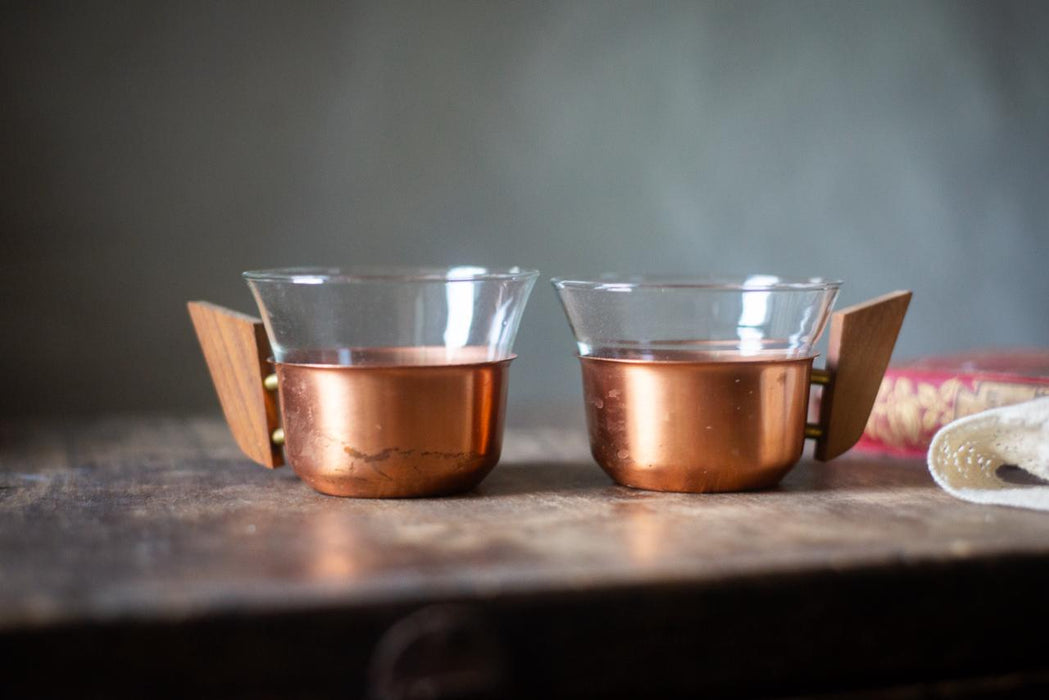 Two copper-colored glasses with wooden handles sit on a wooden surface, with a blurred background.