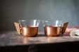 Two copper-colored glasses with wooden handles sit on a wooden surface, with a blurred background.