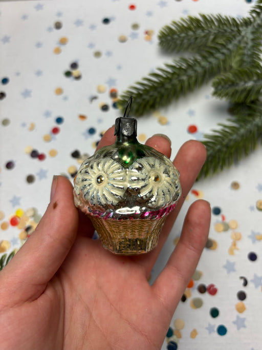 A hand holding a small, ornate glass ornament with a floral design, against a background of confetti and a pine tree branch.