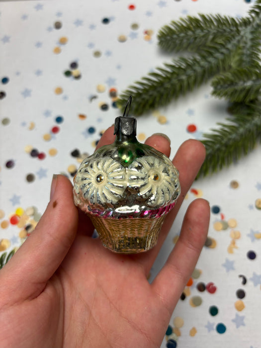 A hand holding a small, ornate glass ornament with a floral design, against a background of confetti and a pine tree branch.