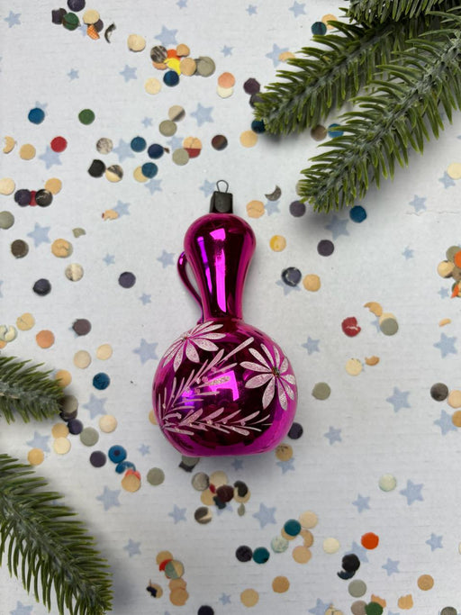 A pink glass ornament with a floral design is surrounded by confetti and greenery on a white background.