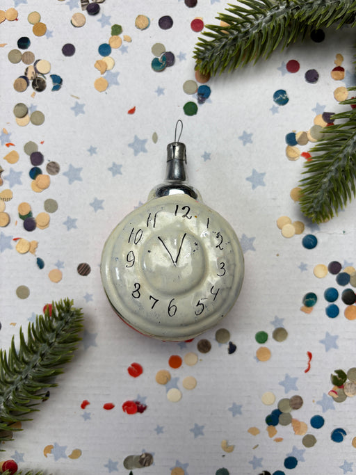 A white ornament with black numbers and hands is surrounded by colorful confetti and pine branches on a white background.