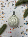 A white ornament with black numbers and hands is surrounded by colorful confetti and pine branches on a white background.