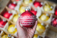A hand holding a red ornament with white stripes against a background of various other red and yellow Christmas ornaments.