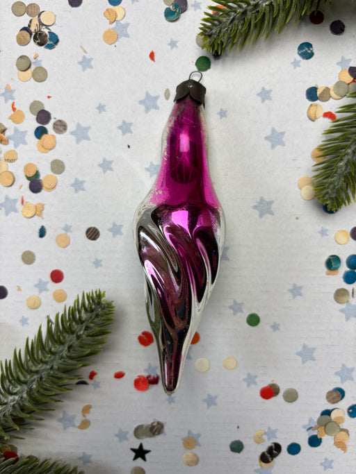 A purple glass ornament with a swirly design is placed on a white background, surrounded by scattered confetti and pine branches.