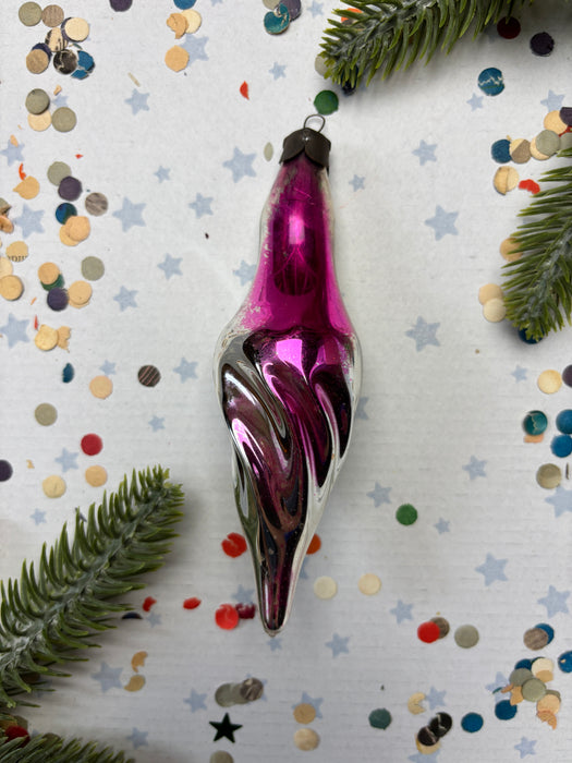 A purple glass ornament with a swirly design is placed on a white background, surrounded by scattered confetti and pine branches.