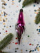 A purple glass ornament with a swirly design is placed on a white background, surrounded by scattered confetti and pine branches.