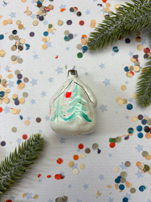 A white Christmas ornament with a green tree design is surrounded by colorful confetti and pine branches on a white background.