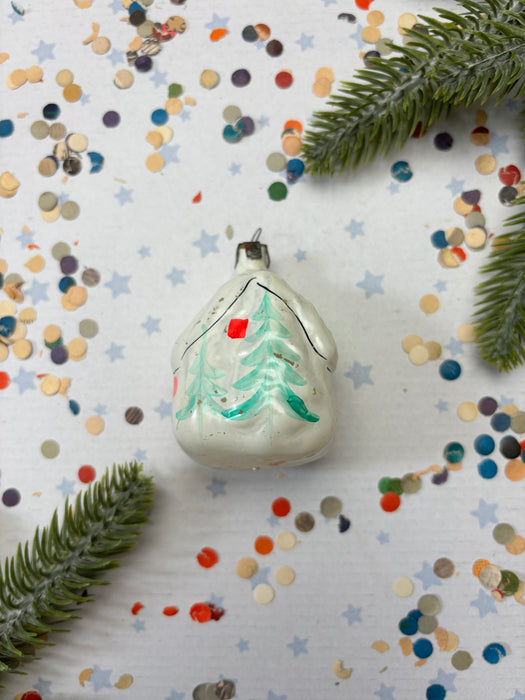 A white Christmas ornament with a green tree design is surrounded by colorful confetti and pine branches on a white background.
