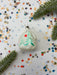 A white Christmas ornament with a green tree design is surrounded by colorful confetti and pine branches on a white background.