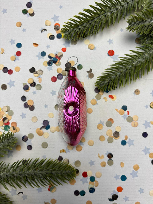 A pink glass ornament with a star design sits on a background of colorful confetti and green pine branches.