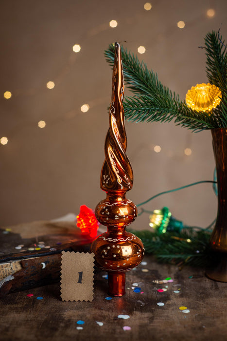 a Christmas tree ornament with a twisted design, surrounded by holiday decorations such as a pine branch, twinkling lights, and confetti on a wooden surface.