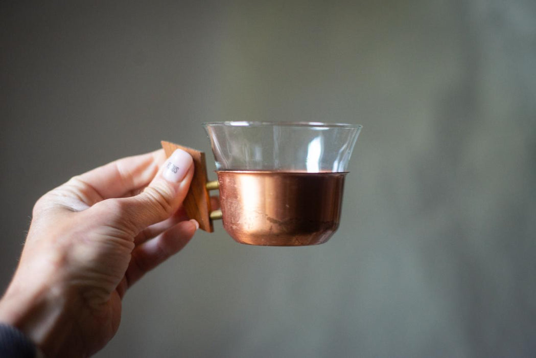 A hand holding a copper-colored glass cup with a wooden handle.