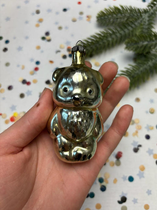 A hand holding a shiny, metallic, bear-shaped ornament against a background of confetti and a pine tree branch.