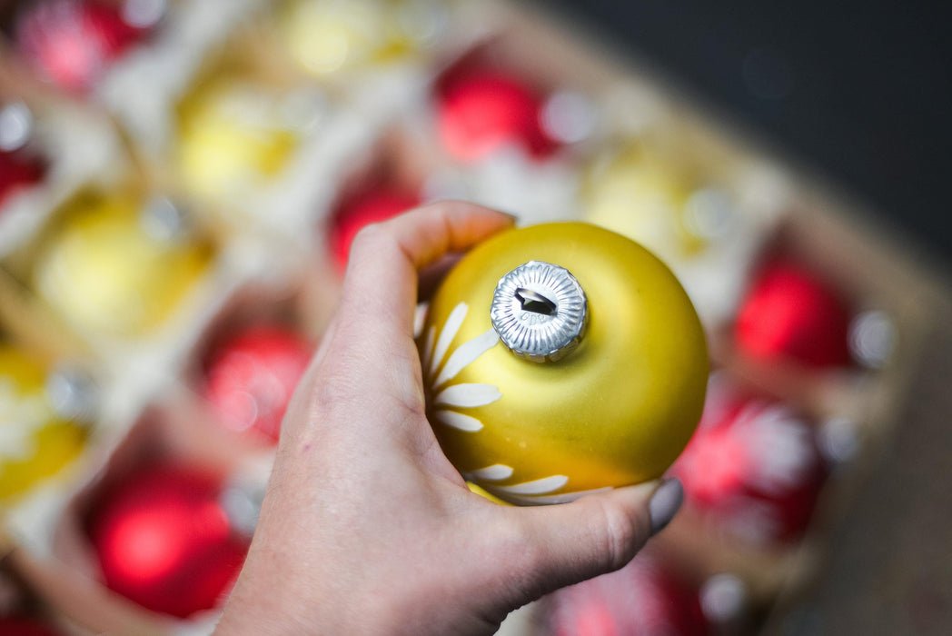 A hand holding a yellow ornament with a silver cap, surrounded by a blurred background of red and yellow ornaments.