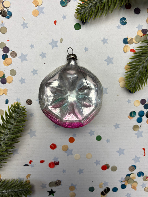 A vintage glass ornament with a pink center is displayed on a white background, surrounded by scattered confetti and pine branches.