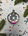 A vintage glass ornament with a pink center is displayed on a white background, surrounded by scattered confetti and pine branches.