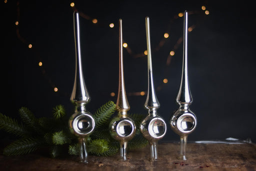 four ornate Christmas ornaments with pointed tops, arranged on a wooden surface against a dark background with twinkling lights.