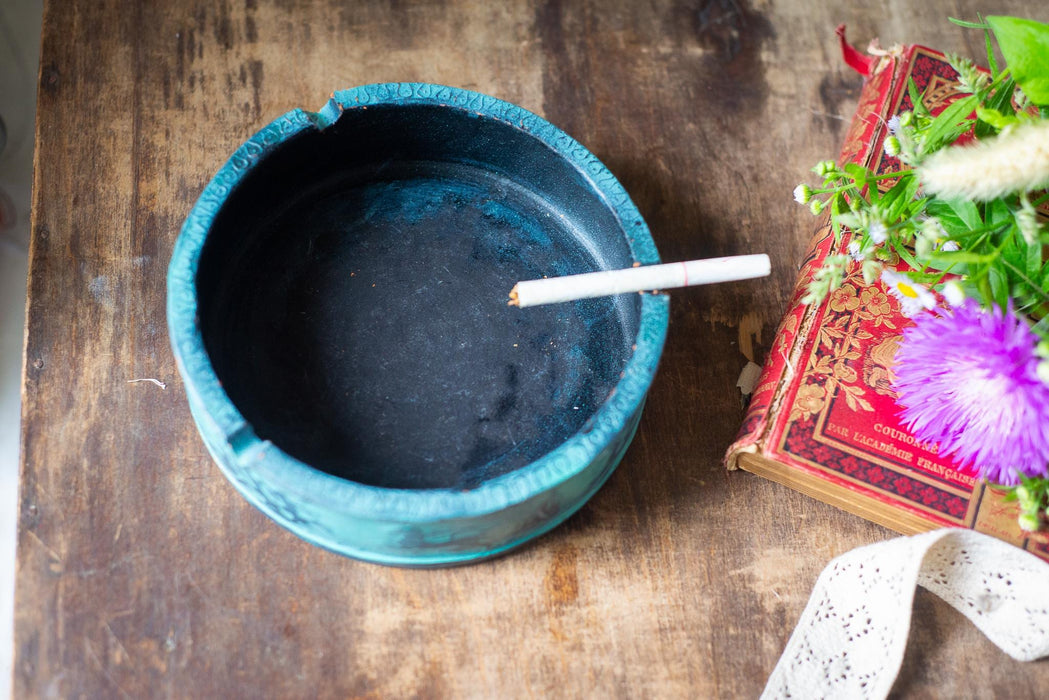 A blue ashtray with a lit cigarette rests on a wooden surface, accompanied by a red book and purple flowers.