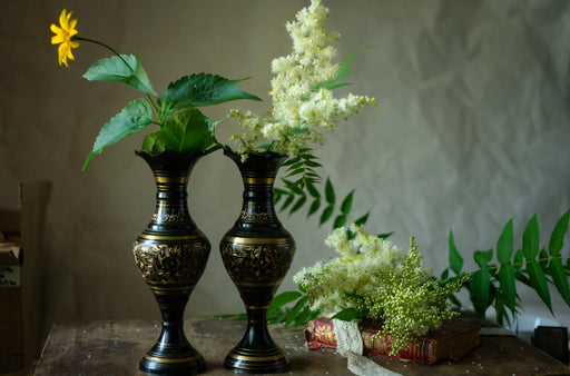 two black vases with gold designs, each containing a single flower, placed on a wooden surface. There are also some green leaves and a book visible in the background.