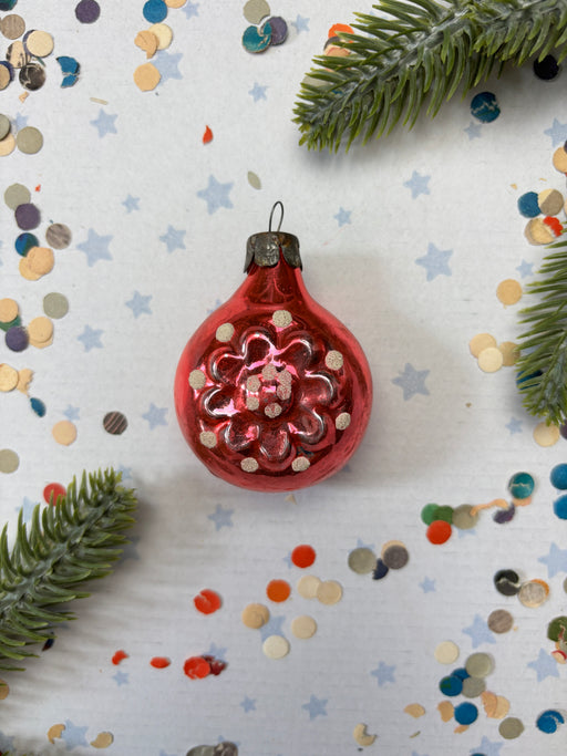 A red Christmas ornament with white flowers is surrounded by scattered confetti and green pine branches on a white background.
