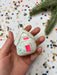 A hand holding a white ornament with a pink and green design, against a background of colorful confetti.