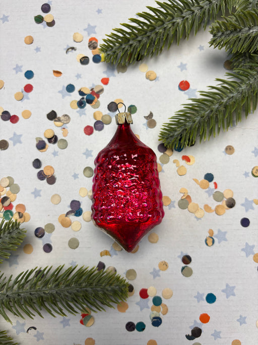 A red glass ornament with a shiny surface is surrounded by colorful confetti and green pine branches on a white background.