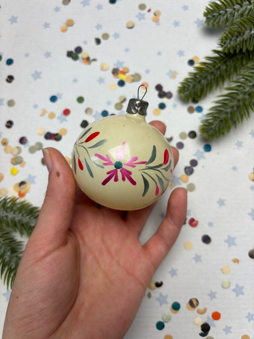 A hand holding a decorative ornament with a floral design against a background of scattered confetti and pine branches.