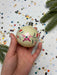 A hand holding a decorative ornament with a floral design against a background of scattered confetti and pine branches.